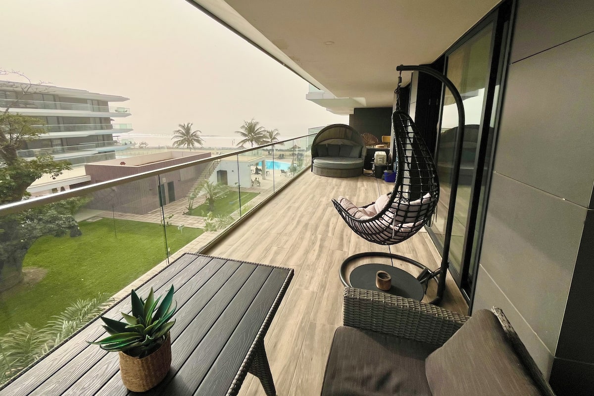 Un balcón con una hamaca colgante, una mesa pequeña con una planta y vistas a una piscina y palmeras.