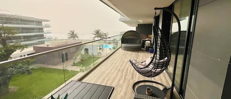A balcony with a hanging chair, a small table with a plant, and a view of a pool and palm trees.