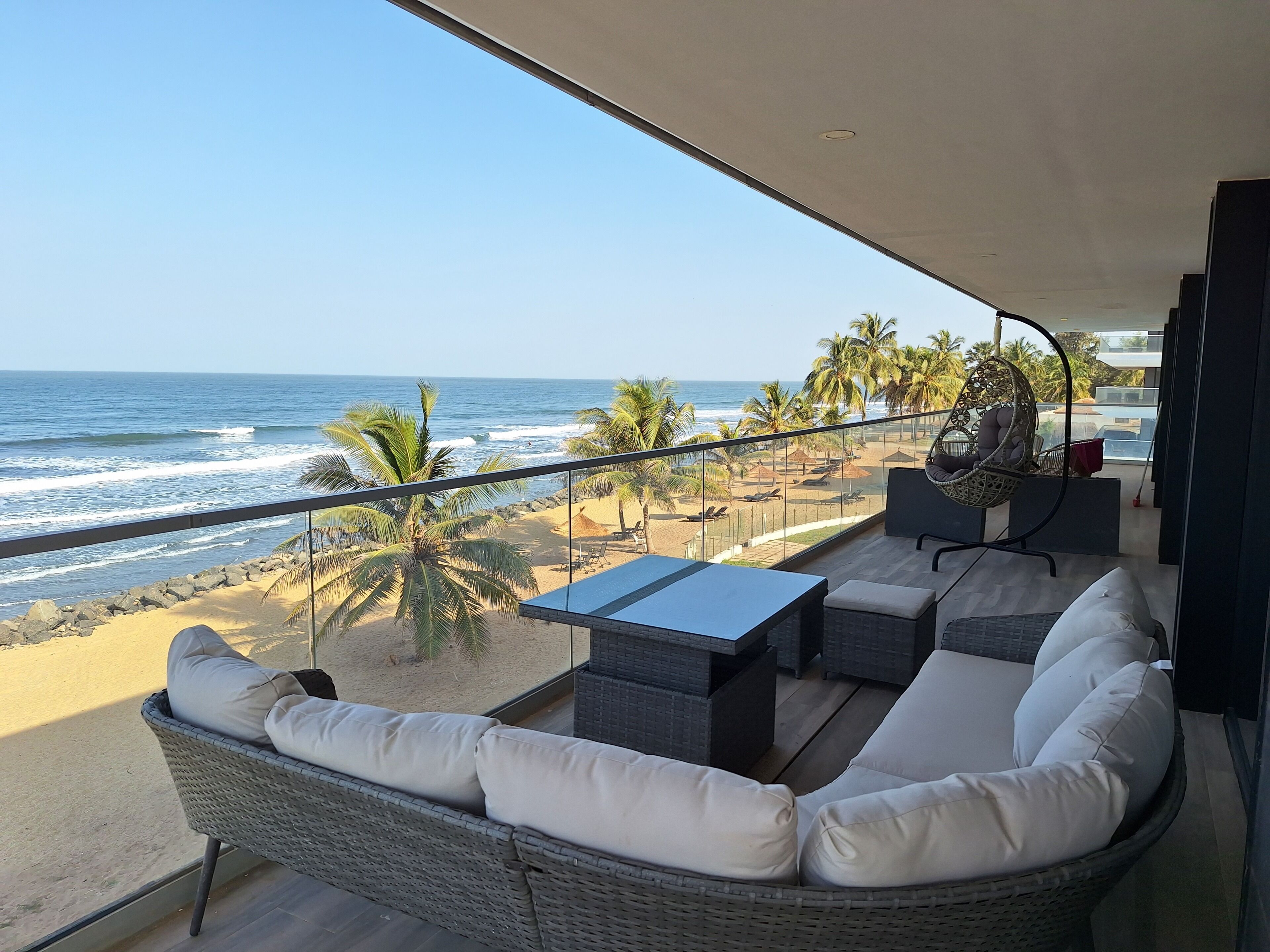 A balcony with a view of the beach and ocean, wicker furniture, and a hanging chair.