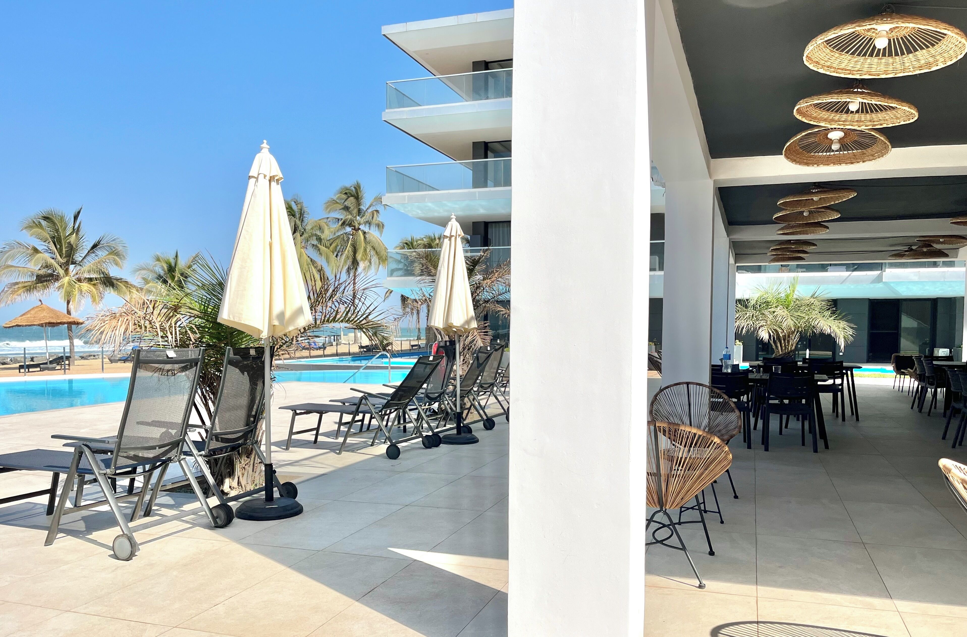 A poolside area with lounge chairs and umbrellas, a dining area with tables and chairs, and a modern building with balconies and outdoor lighting.