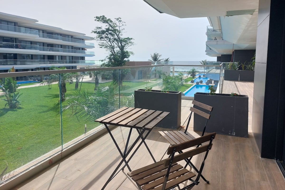 Un balcón con una mesa y sillas de madera, con vistas a una piscina y un césped cuidado.