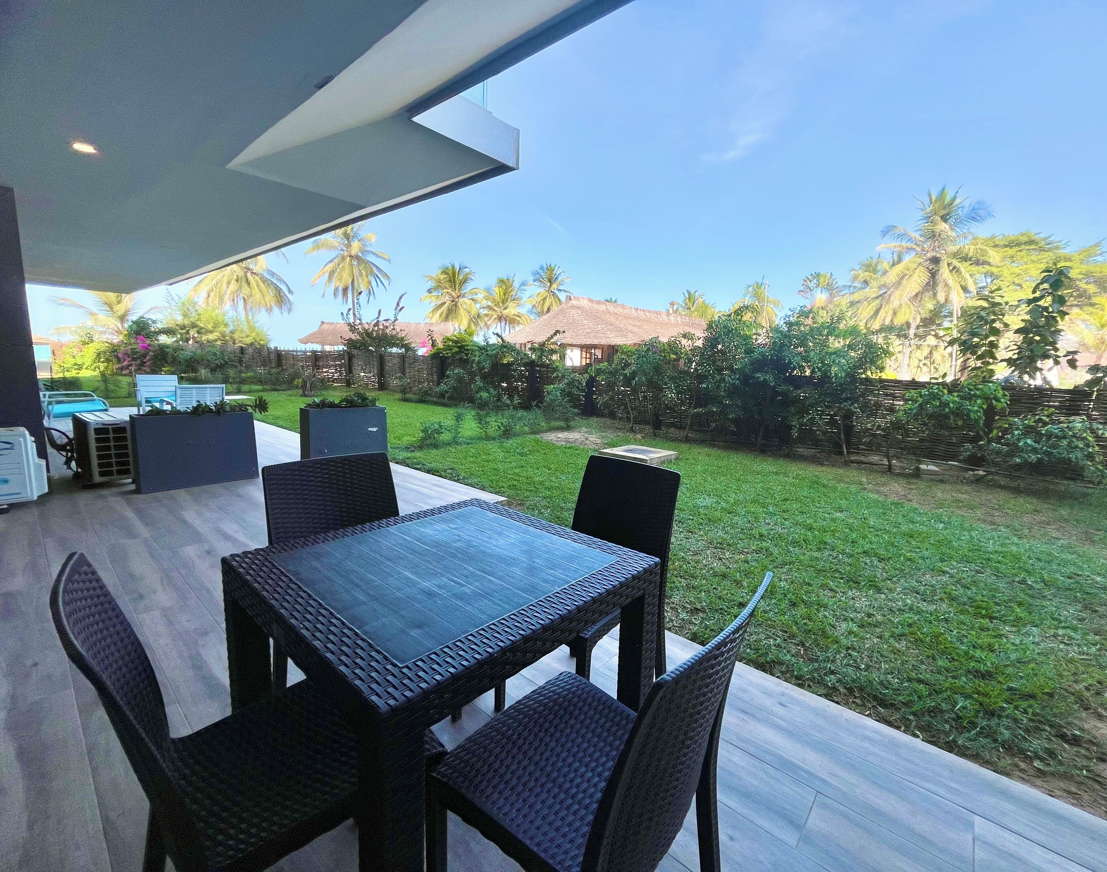 An outdoor patio with a table and chairs, a pool, and a view of a garden with palm trees.