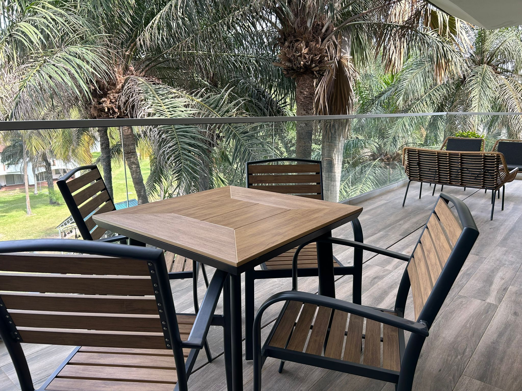 A balcony with a wooden table and chairs, surrounded by lush greenery and palm trees.