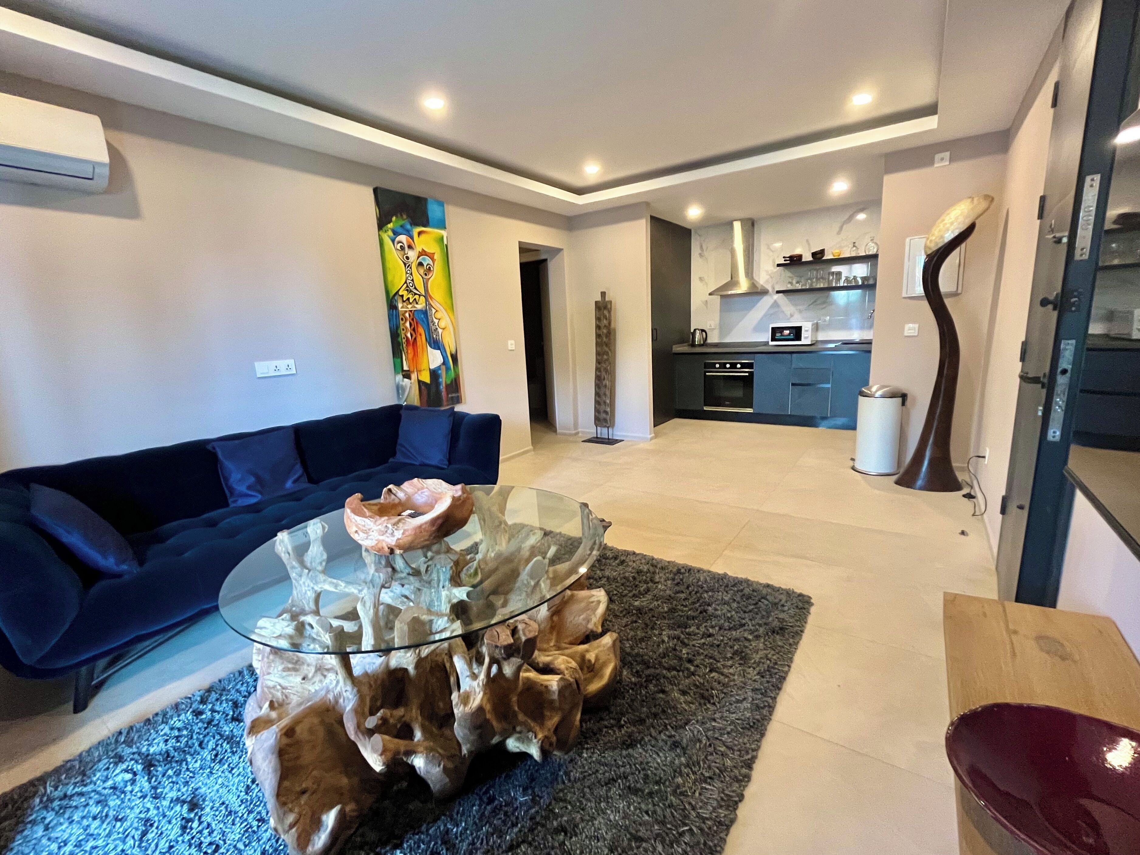 Een moderne woonkamer met een glazen salontafel, een blauwe bank en een keuken op de achtergrond.