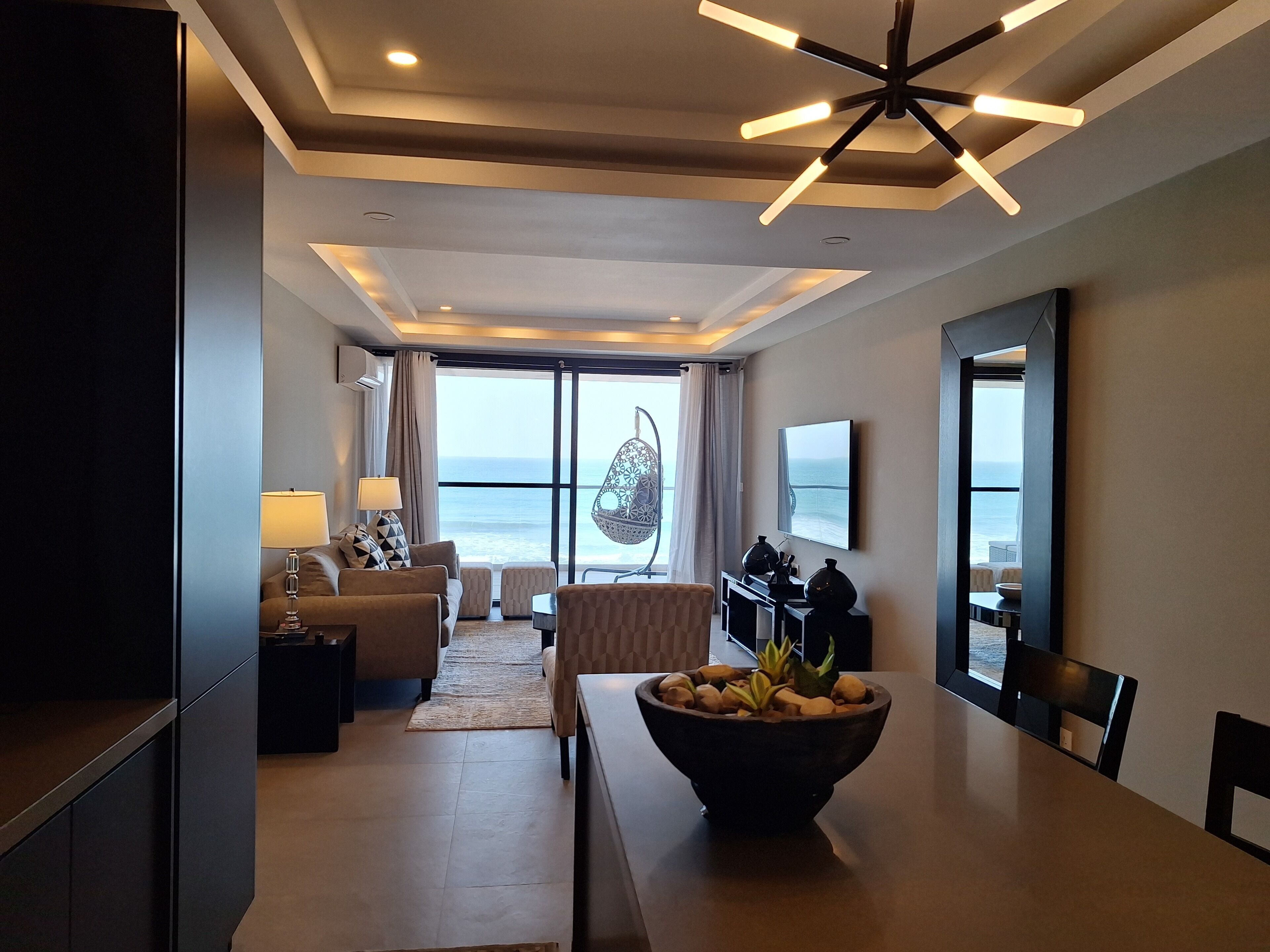 Une chambre d’hôtel moderne dotée d’un coin repas, d’une grande fenêtre donnant sur la mer, d’un fauteuil suspendu et d’un téléviseur à écran plat.