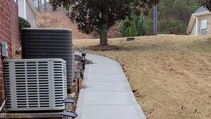 A concrete pathway bordered by a grassy area, with outdoor lighting fixtures and an air conditioning unit visible. - Two Bedroom Basement Apartment (Conyers)