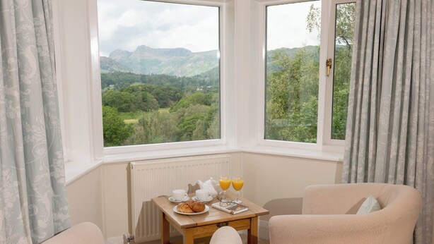 Una habitación con dos ventanas que dan a un paisaje montañoso, una mesa de madera con alimentos para el desayuno y dos sillas color beige.