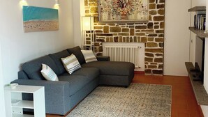 A living room with a grey sofa, a white radiator, a stone wall, and a painting above the radiator. - HOUSE OLEANDRI 200m from the beach (PUNTA ALA)