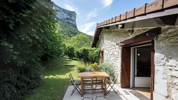 A rustic stone building with a wooden table and chairs in the foreground, a stone wall, and a view of a green valley with a cliff in the background.