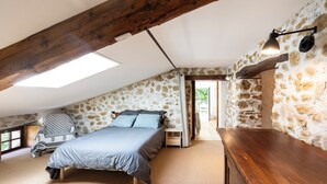 A bedroom with a sloped ceiling, stone walls, a bed with a grey bedspread, a wooden nightstand, and a hanging light fixture.