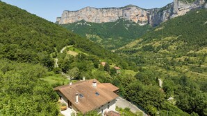 A rural house with a sloped roof, surrounded by lush greenery and a winding road.