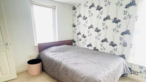 A bedroom with a bed, a window, a radiator, and a wall with floral wallpaper. - Immaculate 3-bed House in Waltham Cross (Waltham Cross)