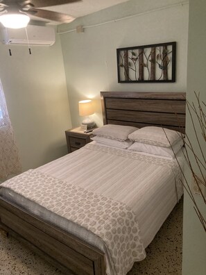 A bedroom with a wooden bed, a nightstand with a lamp, a wall-mounted air conditioner, and a framed artwork above the bed.
