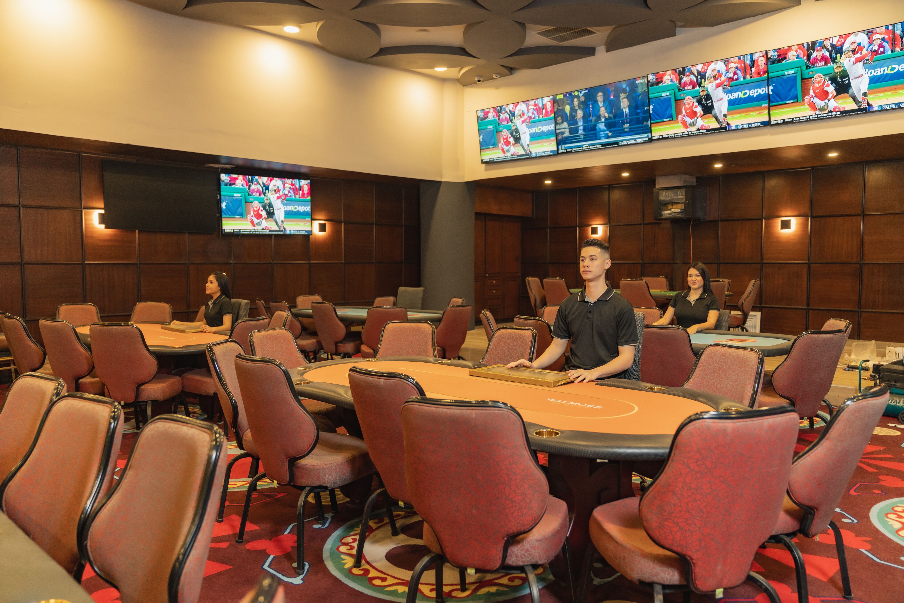 Un salón de casino con múltiples mesas de blackjack, sillas y una pantalla grande que muestra eventos deportivos.
