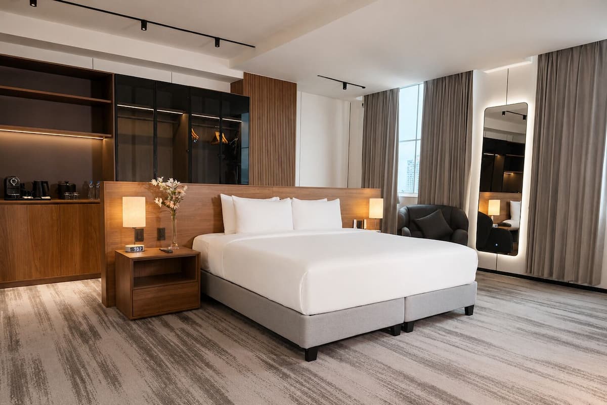 A modern hotel room with a large bed, a wooden nightstand, a desk with a lamp, and a mirror.