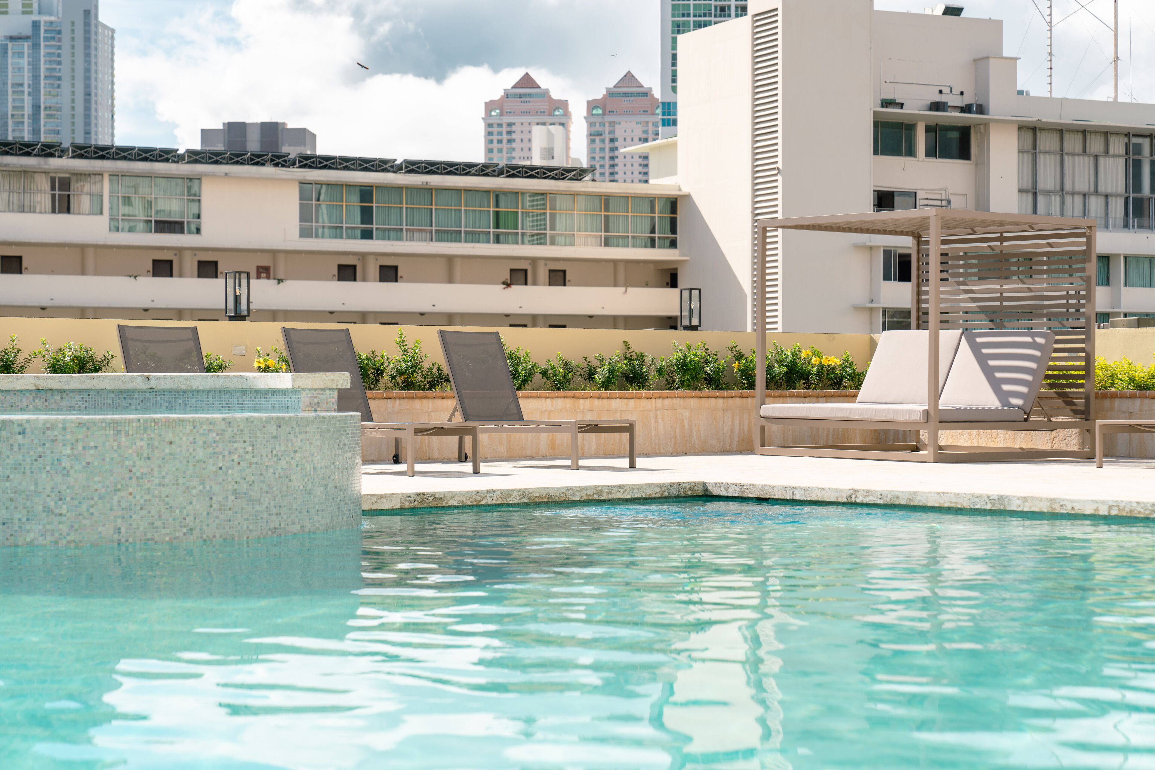 Una piscina con sillones de descanso y una pérgola, rodeada por un muro bajo y un paisaje urbano de fondo.