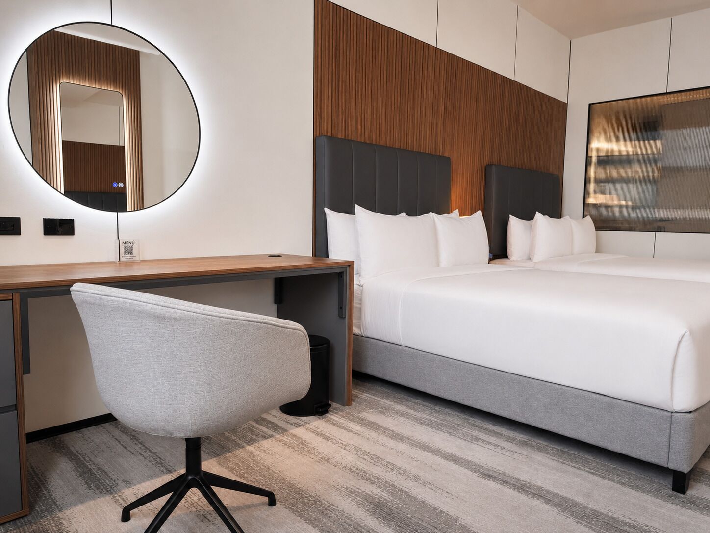 Une chambre d’hôtel équipée d’un lit, d’un bureau, d’une chaise, d’un miroir rond et d’un téléviseur fixé au mur.