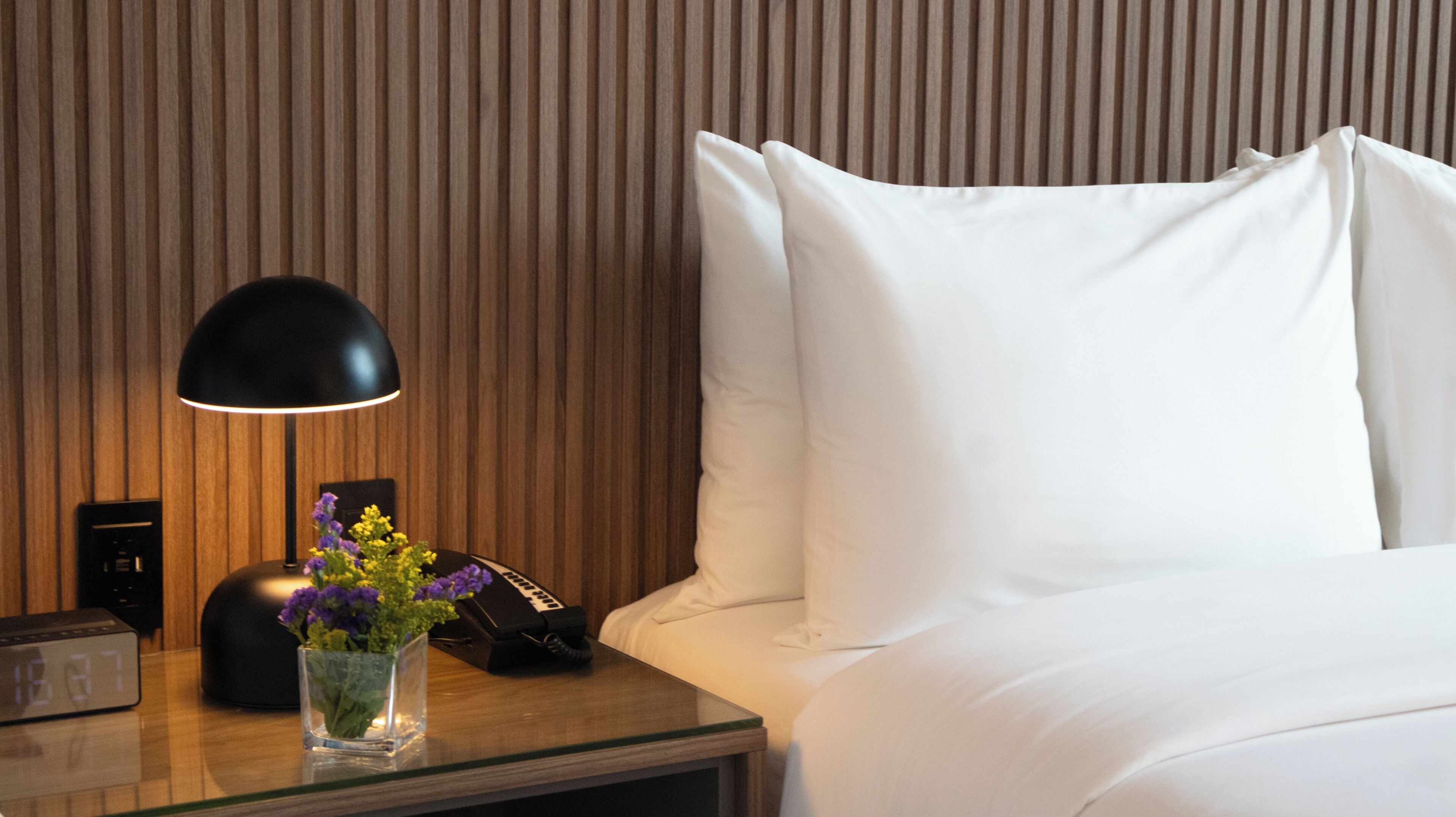 A hotel room with a wooden panel wall, a bedside table with a lamp, a vase with flowers, a telephone, and a bed with white bedding.