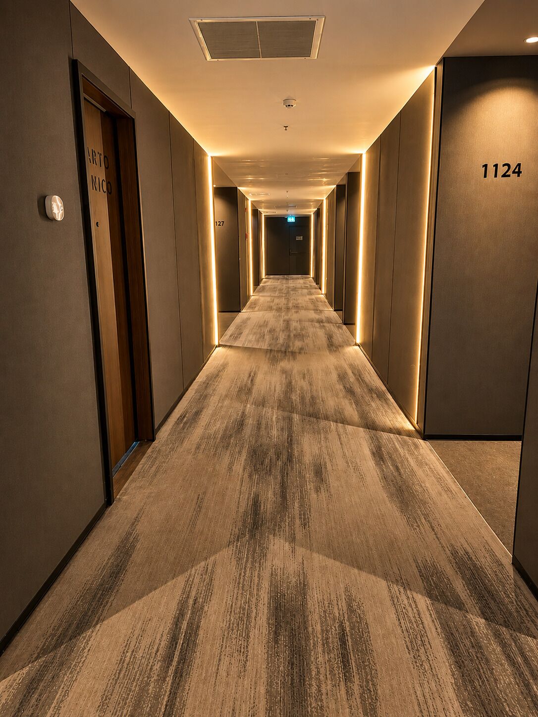 Ein Hotelkorridor mit Teppichböden, Türen mit den Nummern 1124 und 127 sowie einer Decke mit einer Leuchte.