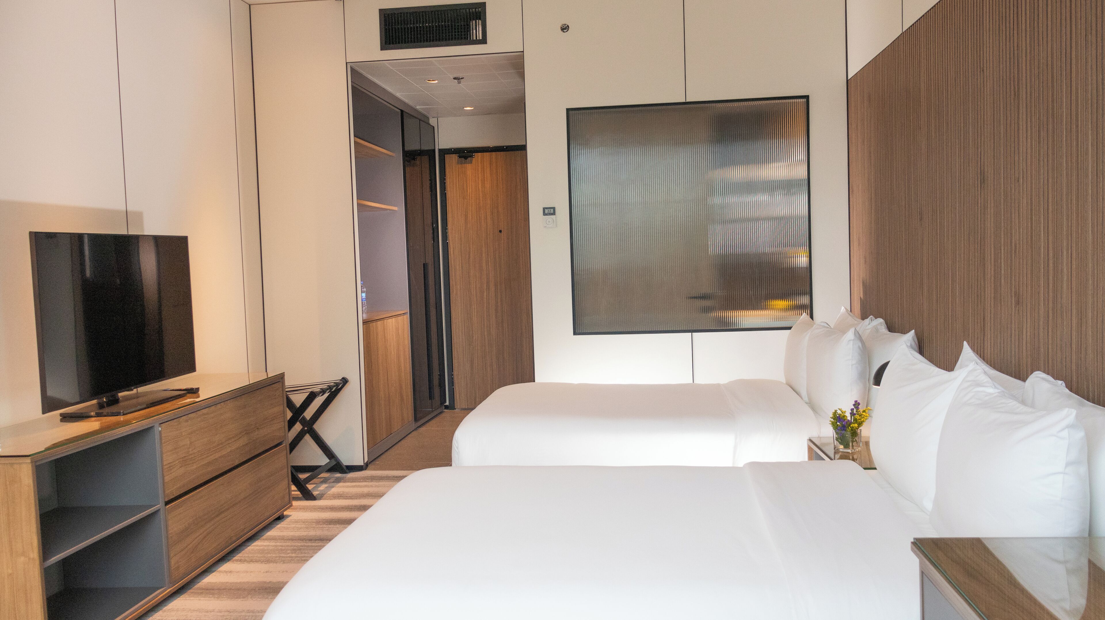 A modern hotel room with a large flat-screen TV, a wooden dresser, a bed with white linens, and a large window with a view.