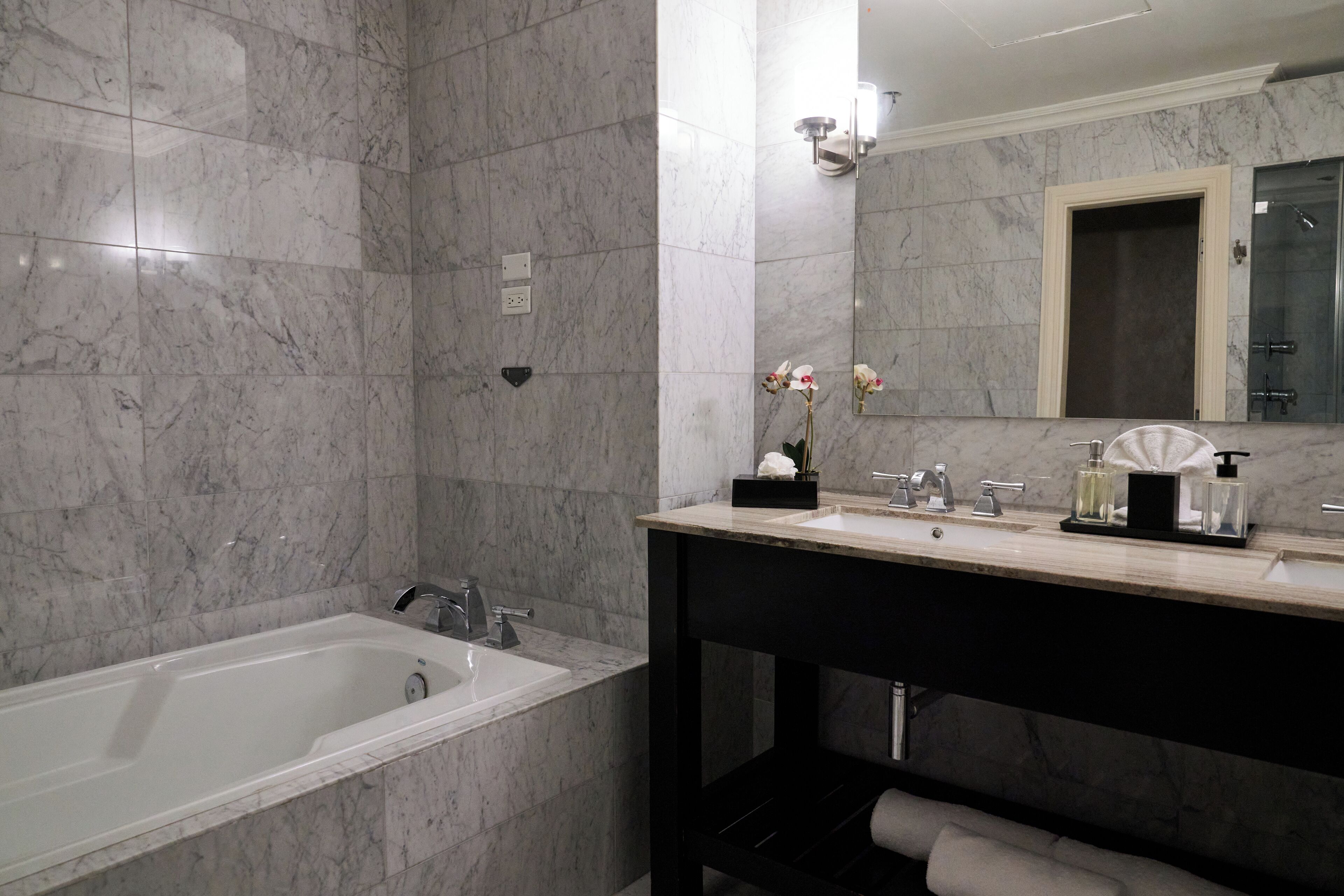 Ein modernes Badezimmer mit Marmorwand, einer Badewanne, einem Waschbecken inklusive Spiegel und einem Handtuchhalter.