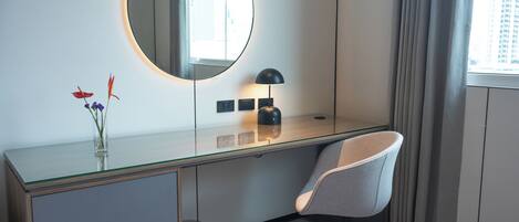 Una habitación de hotel moderna con un escritorio de vidrio, un espejo redondo, una silla gris y un pequeño jarrón con flores.