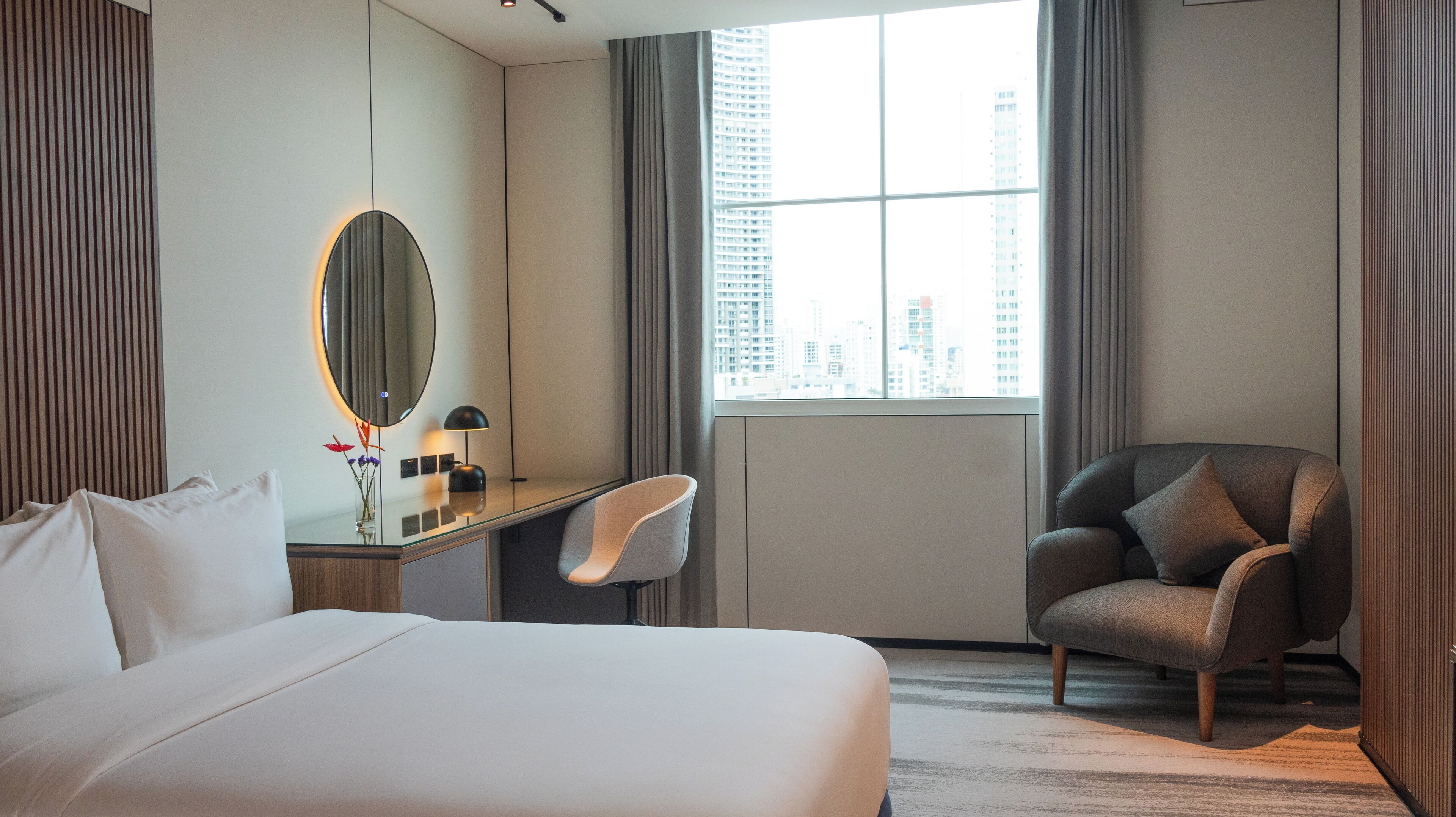 A hotel room with a bed, a desk with a chair, a round mirror, and a window with a city view.