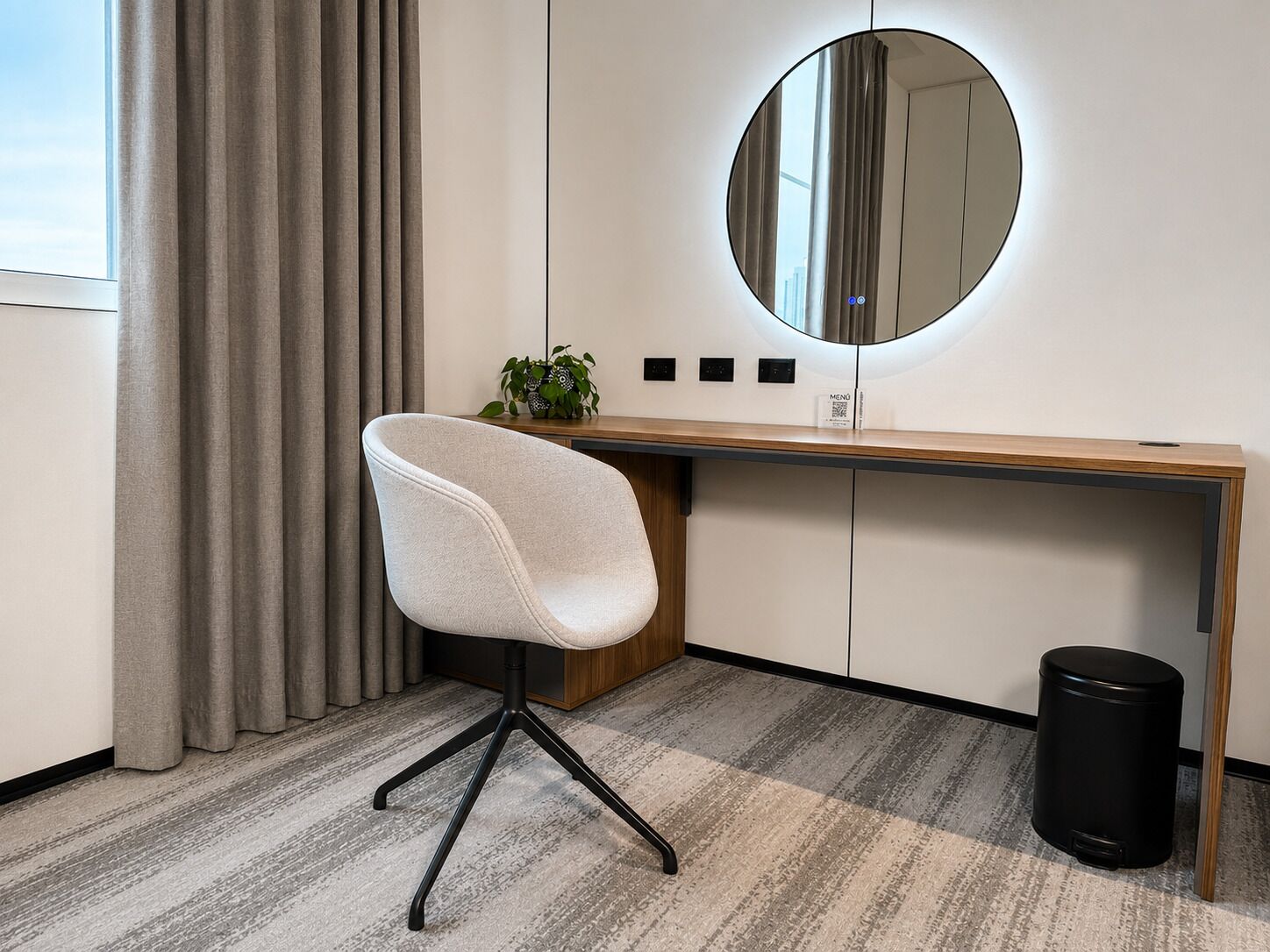 A modern office space with a desk, chair, mirror, potted plant, and trash bin.