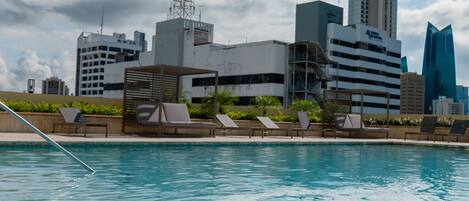 Una piscina con sillones de descanso y un horizonte urbano al fondo.