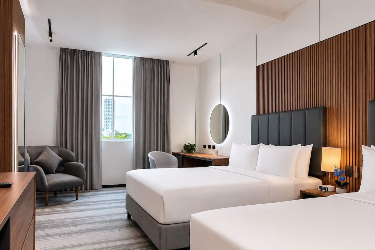A hotel room with two beds, a desk, a chair, a large window with curtains, and a view of buildings.