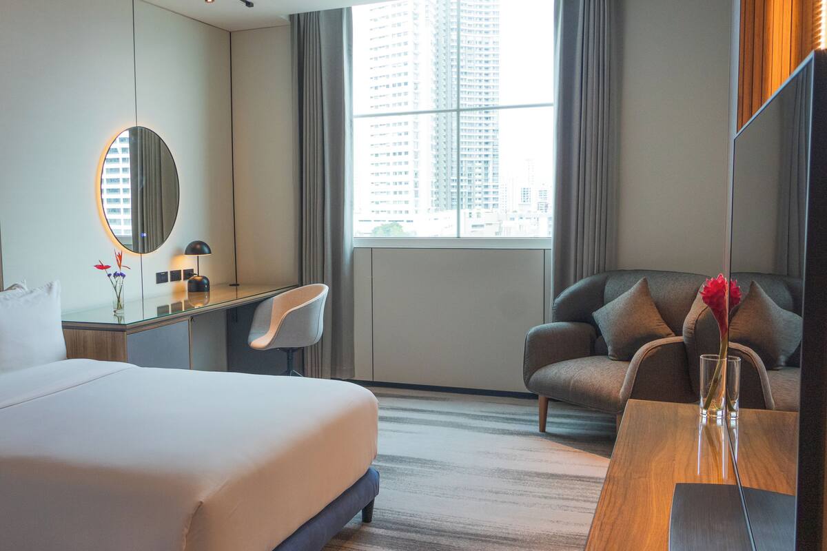 A hotel room with a large bed, a desk, a chair, a mirror, and a view of buildings through the window.