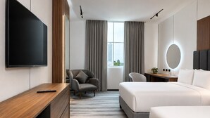 Habitación de hotel moderna con televisor de pantalla plana grande, escritorio, sillón cómodo, cama, mesita de noche y vistas a la ciudad desde la ventana.