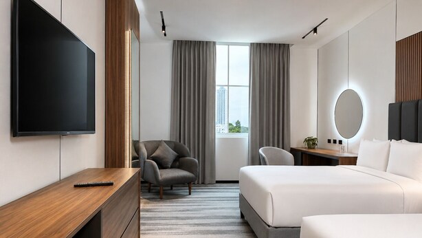 Habitación de hotel moderna con televisor de pantalla plana grande, escritorio, sillón cómodo, cama, mesita de noche y vistas a la ciudad desde la ventana.