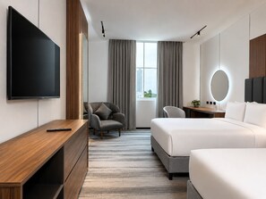 Habitación de hotel moderna con televisor de pantalla plana grande, escritorio, sillón cómodo, cama, mesita de noche y vistas a la ciudad desde la ventana.