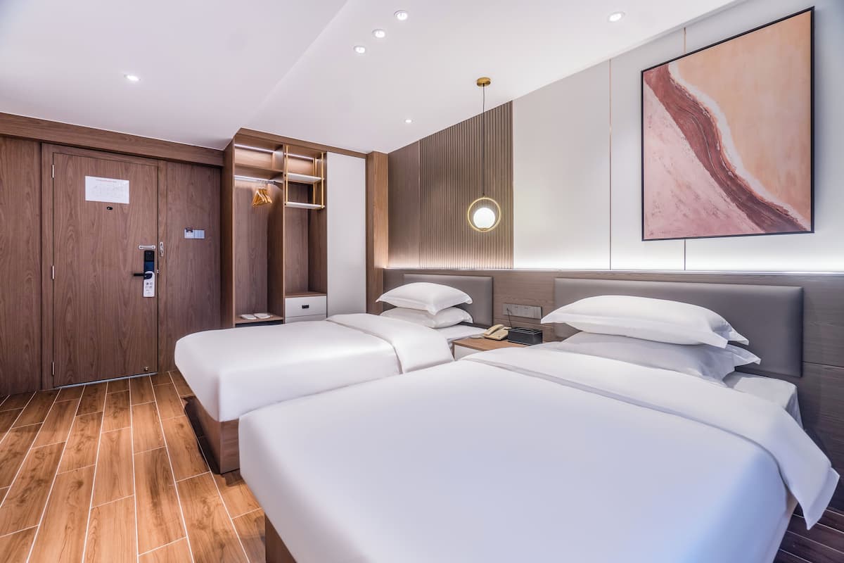 A modern hotel room with two beds, a wardrobe, a bedside table with a lamp, and a large abstract painting on the wall.