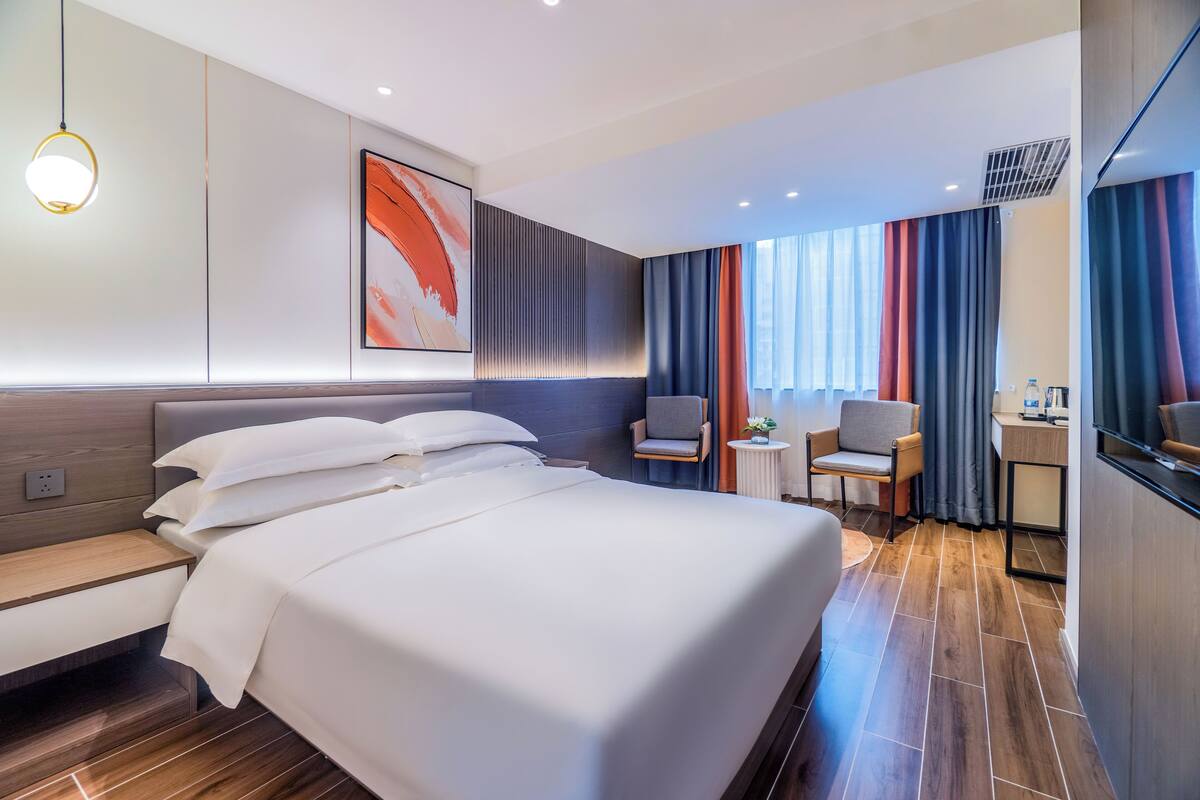 A modern hotel room with a large bed, a desk, a chair, and a small table with a plant.