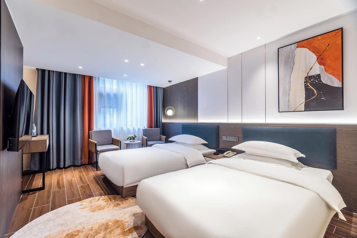 A hotel room with two beds, a desk, a chair, a small table, and a painting on the wall.