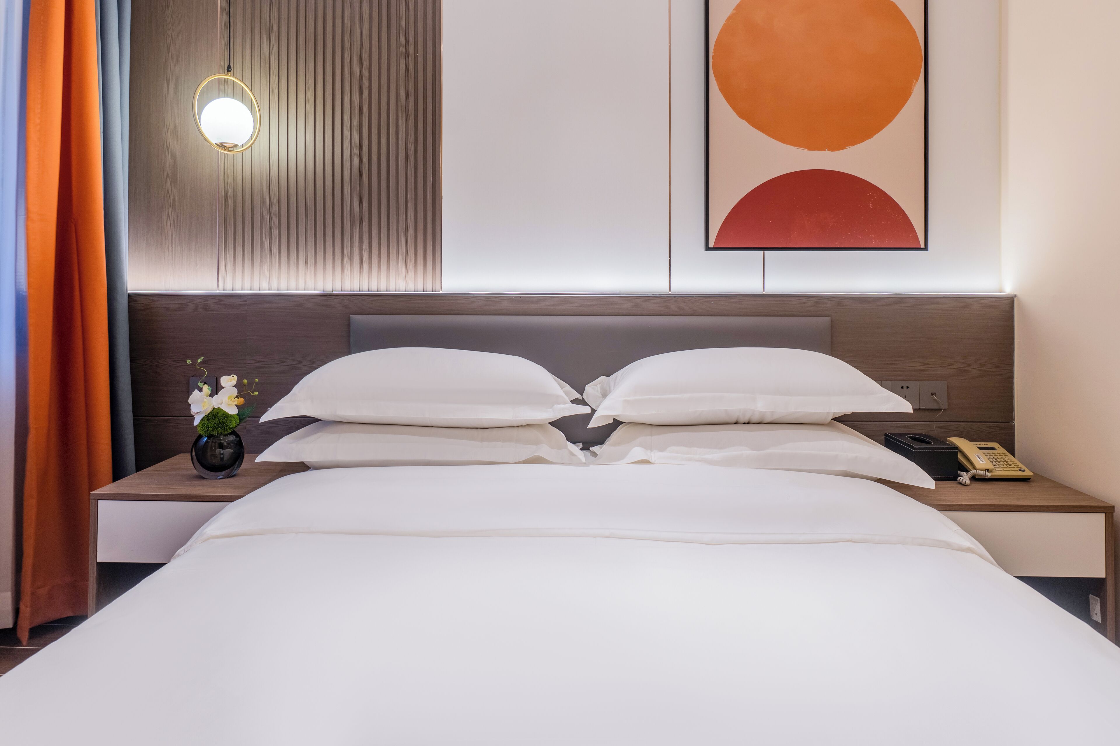 A neatly made bed with white linens, a bedside table with a lamp, a phone, and a vase with flowers.
