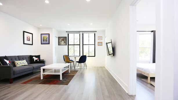 Una sala moderna con un sofá gris, una mesa de centro blanca y dos sillas. Hay ventanas amplias con persianas, un televisor de pantalla plana montado en la pared y una cama al fondo.