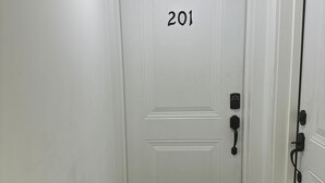 Una puerta blanca con el número 201, un felpudo negro con los mensajes “GOODBYE” y “HELLO!”, y un timbre negro.