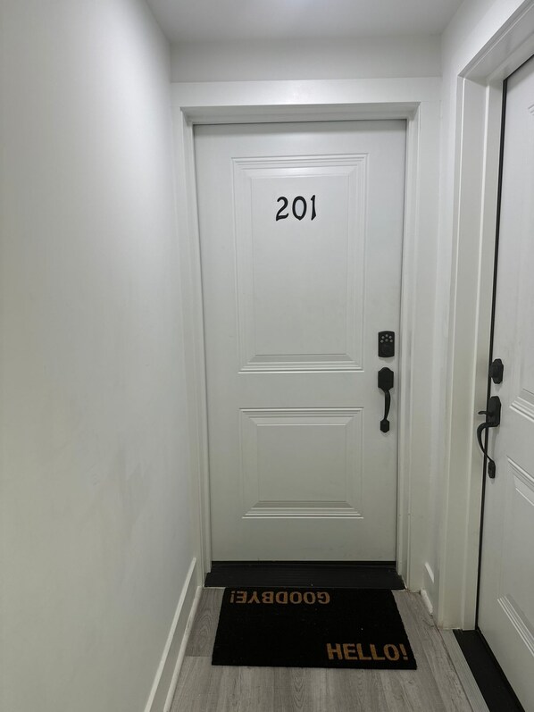 A white door with the number 201, a black doormat with "GOODBYE" and "HELLO!" text, and a black doorbell.
