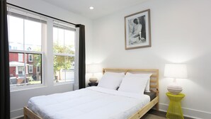 Un dormitorio con cama de madera, ropa de cama blanca, una mesita de noche con lámpara y una ventana con cortinas.
