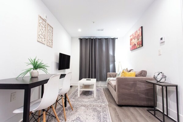 A modern living room with a sofa, a dining table, chairs, a TV, and a plant. - Cozy 1BR in Petworth with Fast Wi-Fi & Walk to Shops (Washington)