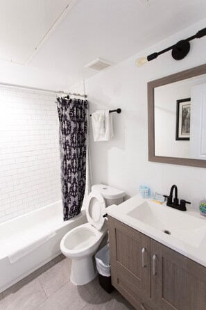 A bathroom with a white toilet, a sink with a mirror above it, and a shower curtain. - Cozy 1BR in Petworth with Fast Wi-Fi & Walk to Shops (Washington)