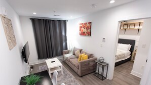 Ett modernt vardagsrum med en soffa, ett litet bord och en tv. Längst bak syns en säng och en dörr som leder till ett annat rum.