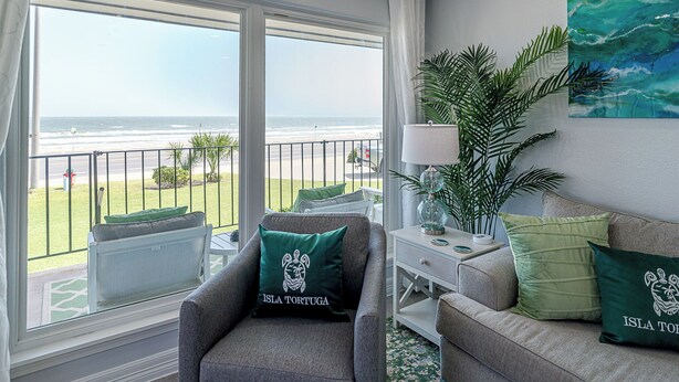 Ein Zimmer mit Blick auf den Strand, eine Couch, ein Sessel, ein Beistelltisch mit einer Lampe und ein Gemälde an der Wand.