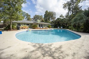 A swimming pool with a sunken center, surrounded by a concrete deck. There is a covered patio area with a table and chairs, and a house in the background.