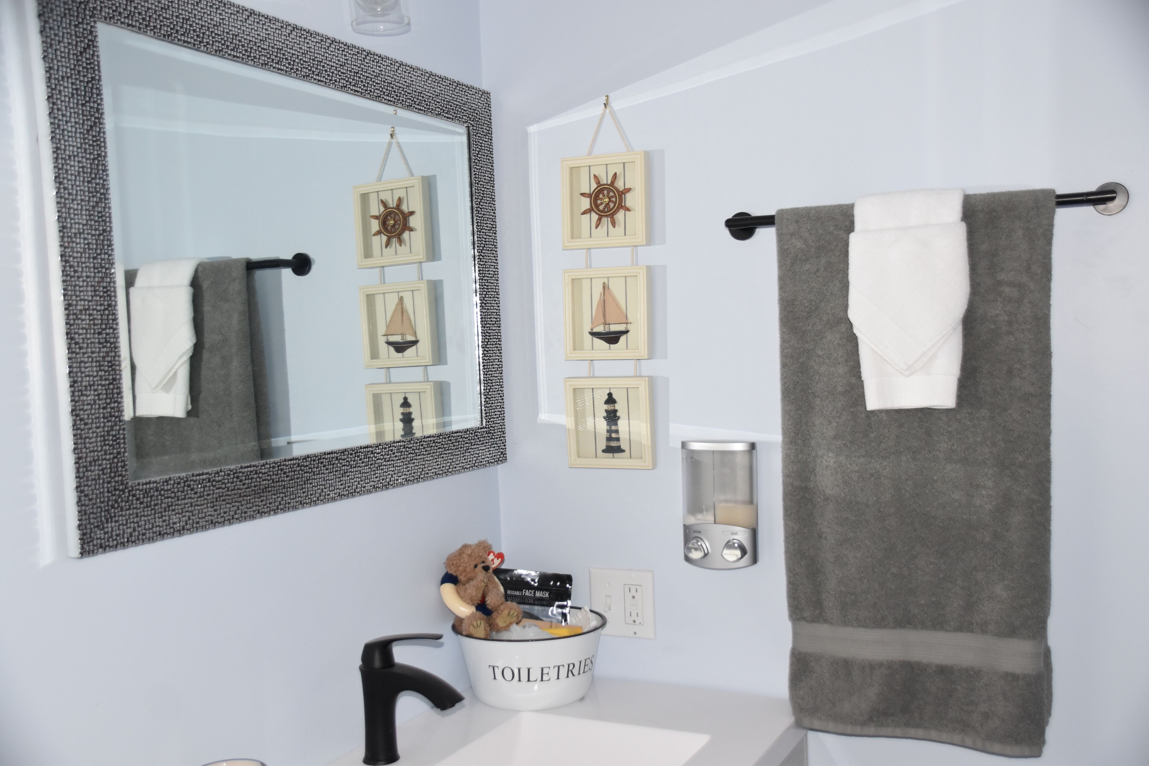 Une salle de bain avec un grand miroir, un porte-serviettes, un lavabo, un distributeur de savon et un ours en peluche dans un récipient.