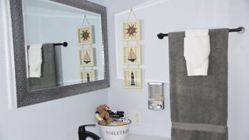 Une salle de bain avec un grand miroir, un porte-serviettes, un lavabo, un distributeur de savon et un ours en peluche dans un récipient.