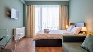 Ein Hotelzimmer mit Bett, Fernseher, Sessel, Tisch und einem Balkon mit Vorhängen.